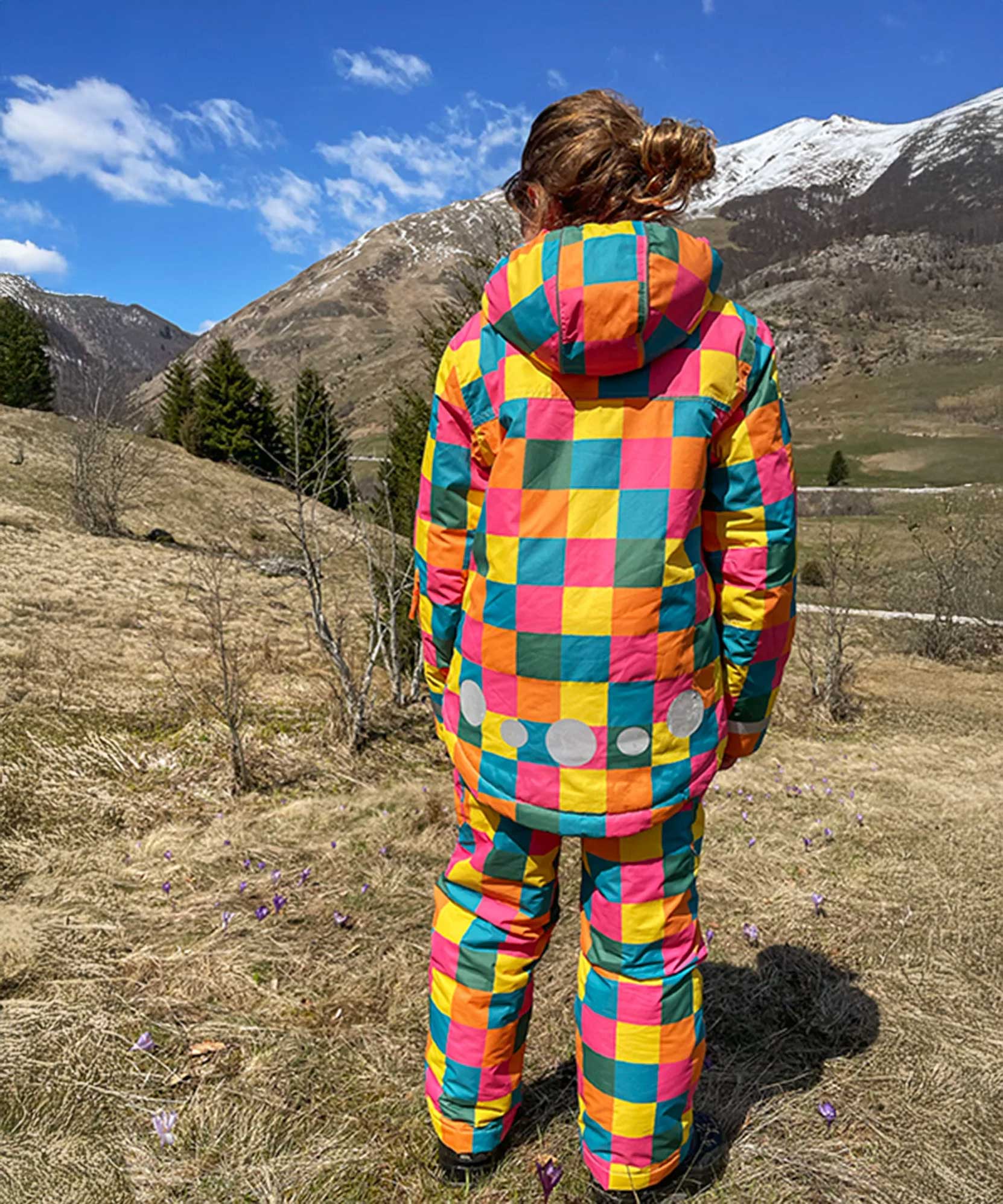 A child facing away from the camera wearing the Frugi Snow and Ski Salopettes in the Rainbow Check design with the matching ski jacket. The design features squares in pink, orange, yellow, blue and green.