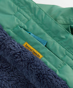A close up of the Frugi kids snow and ski coat in green with a mountain scene print. Showing the thick navy fleece inner and a secret snack pocket on the inside
