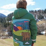 A child facing away from the camera wearing the Frugi Snow And Ski Scene Coat The coat is in a Holly Green colour and features a large Mountains landscape design on the back.
