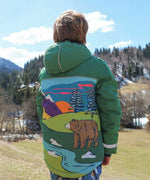 A child facing away from the camera wearing the Frugi Snow And Ski Scene Coat The coat is in a Holly Green colour and features a large Mountains landscape design on the back.
