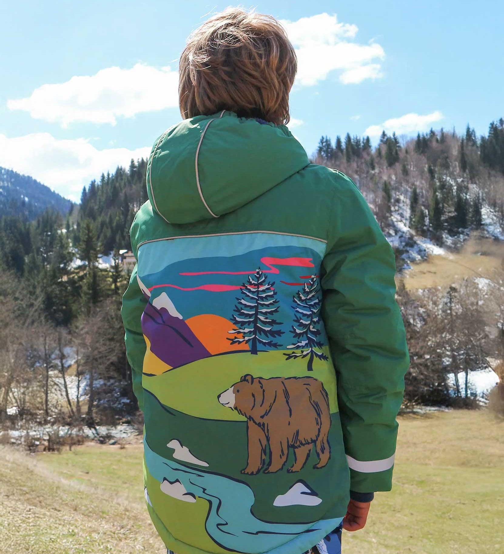 A child facing away from the camera wearing the Frugi Snow And Ski Scene Coat The coat is in a Holly Green colour and features a large Mountains landscape design on the back.
