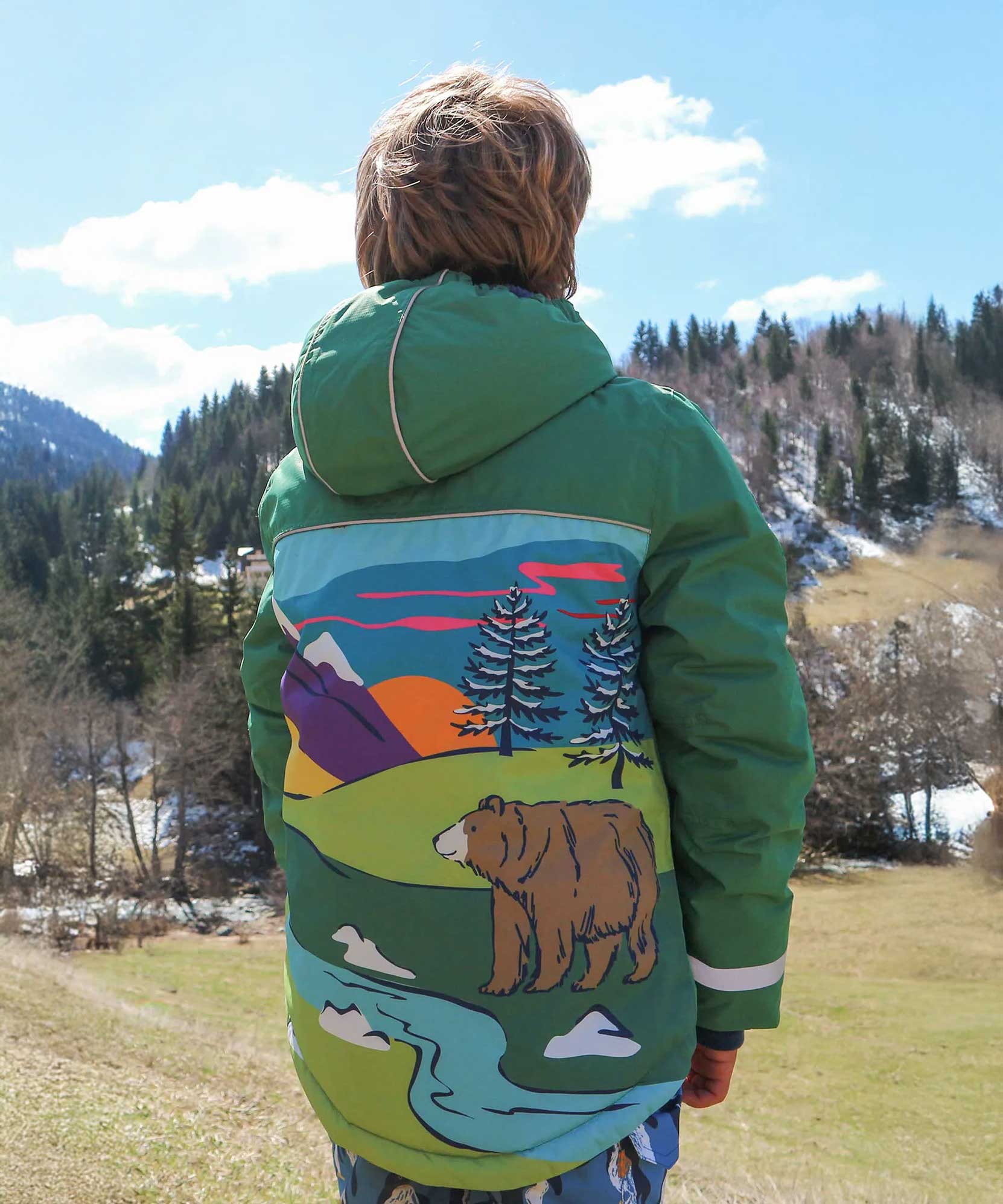 A child facing away from the camera wearing the Frugi Snow And Ski Scene Coat The coat is in a Holly Green colour and features a large Mountains landscape design on the back.
