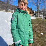 A child wearing the Frugi Snow And Ski Scene Coat in a Holly Green colour with a Mountains design on the back. The child is outdoors and the ground behind then is covered in snow. 