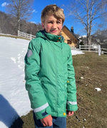 A child wearing the Frugi Snow And Ski Scene Coat in a Holly Green colour with a Mountains design on the back. The child is outdoors and the ground behind then is covered in snow. 