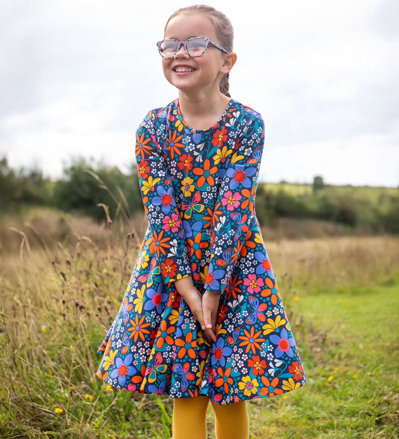 A child wearing a Frugi Organic Cotton Sofia Skater Dress in a Navy with a Retro Floral print with yellow leggings.
