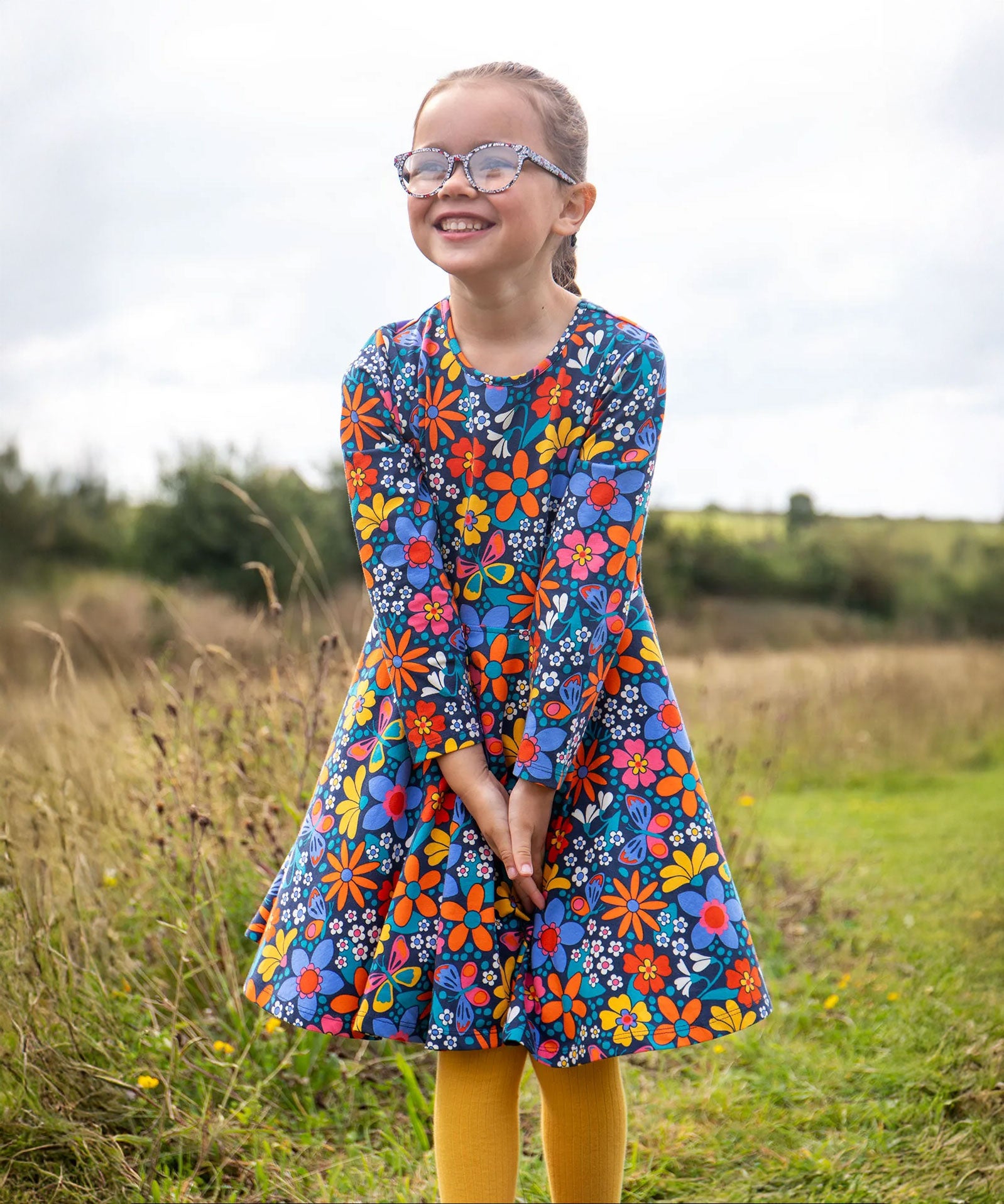 A child wearing a Frugi Organic Cotton Sofia Skater Dress in a Navy with a Retro Floral print with yellow leggings.