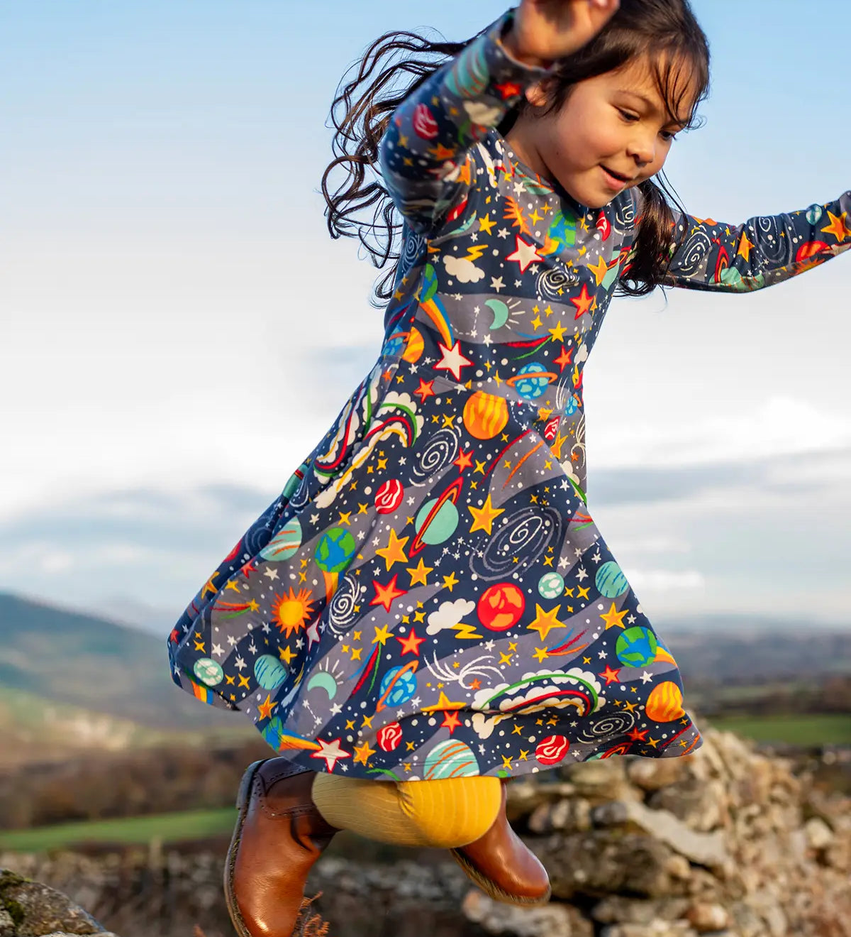 Child wearing the Frugi Sofia organic cotton skater dress in navy with an all over explore the stars print with yellow tights and brown boots. 