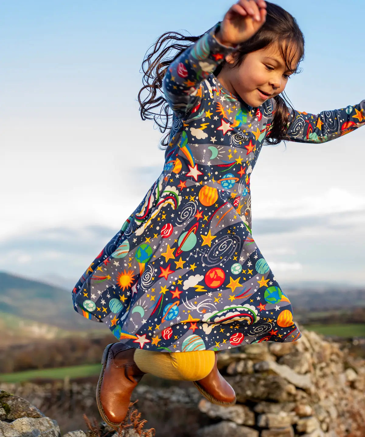 Child wearing the Frugi Sofia organic cotton skater dress in navy with an all over explore the stars print with yellow tights and brown boots. 