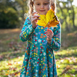A child wearing the Frugi Sofia skater dress with a wild woods print. A beautiful turquoise blue with a pattern all over featuring flowers, greenery and mushrooms.
