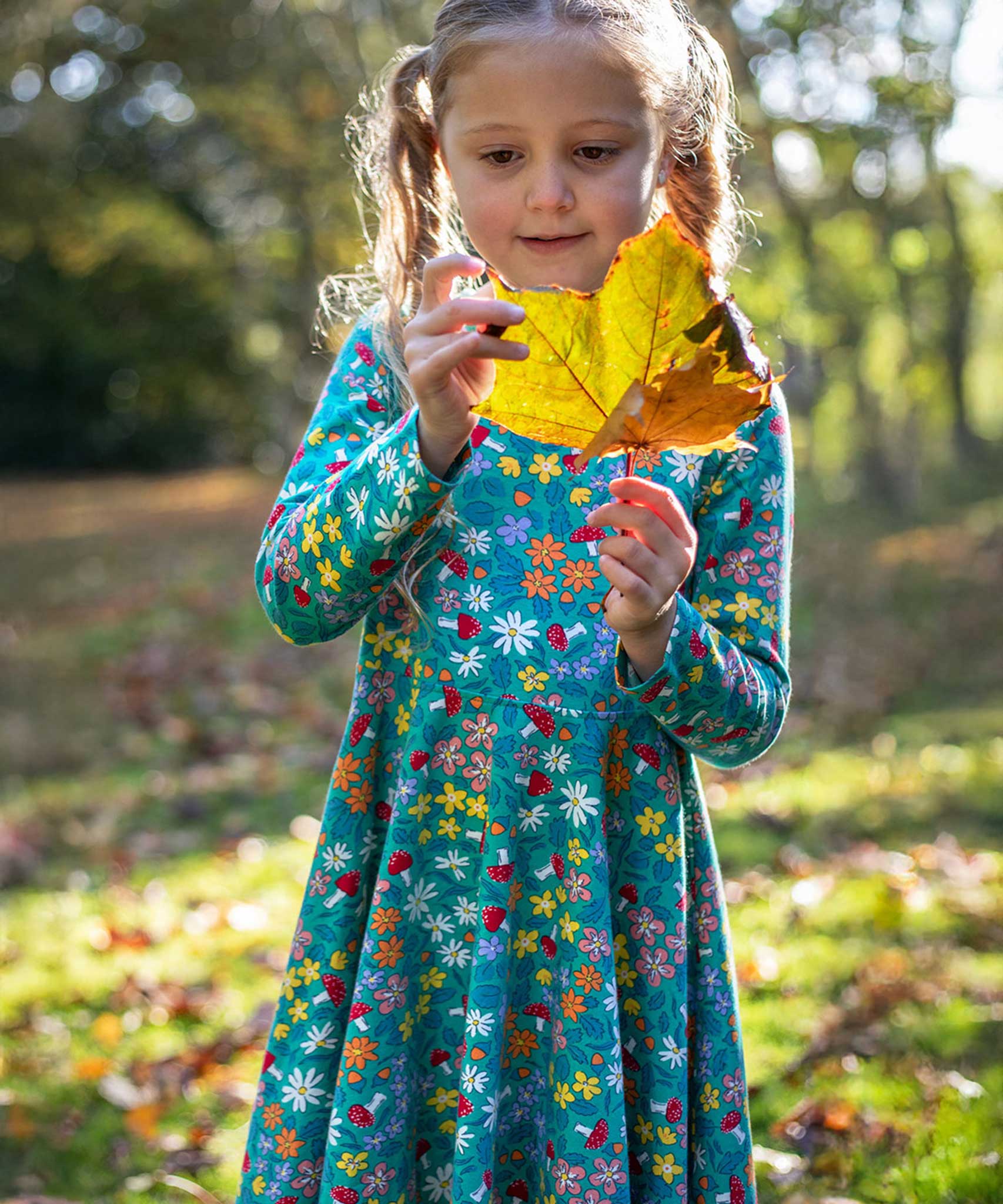 A child wearing the Frugi Sofia skater dress with a wild woods print. A beautiful turquoise blue with a pattern all over featuring flowers, greenery and mushrooms.