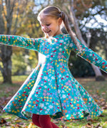 A child wearing the Frugi Sofia skater dress with a wild woods print. A beautiful turquoise blue with a pattern all over featuring flowers, greenery and mushrooms.