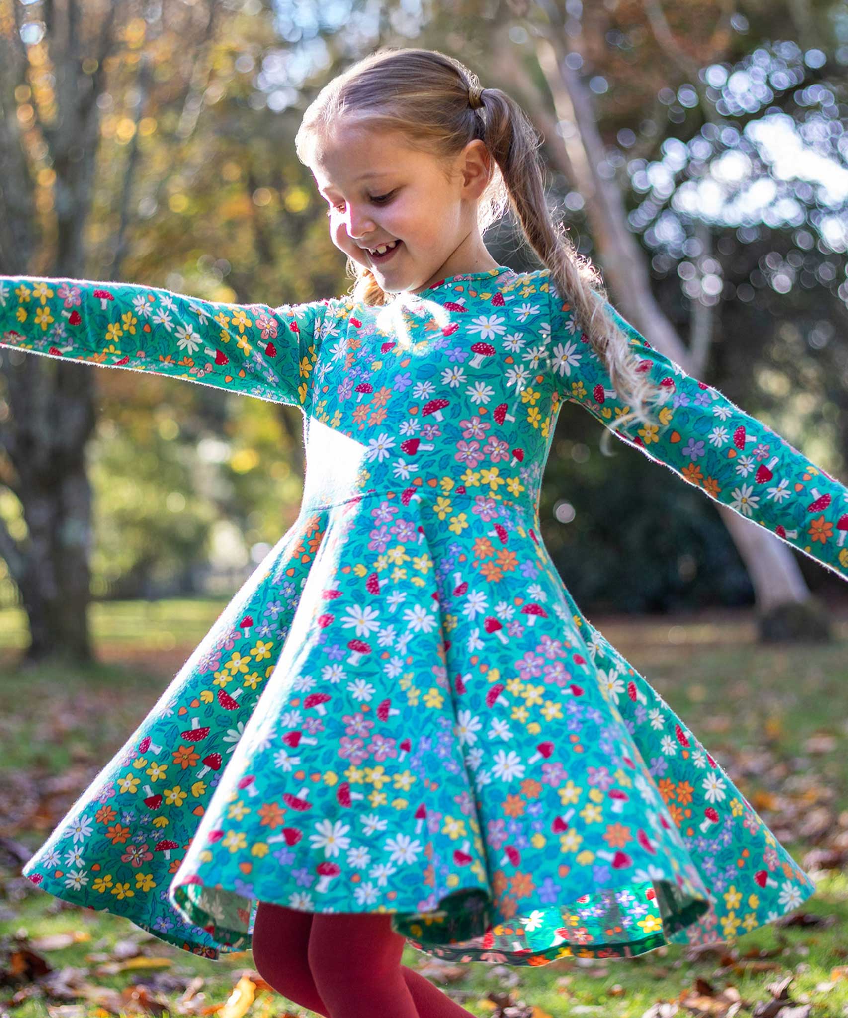 A child wearing the Frugi Sofia skater dress with a wild woods print. A beautiful turquoise blue with a pattern all over featuring flowers, greenery and mushrooms.