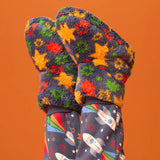 A pair of fuzzy knit mittens with a colorful star pattern in orange, yellow, green, and red on a dark gray background, shown on a child's hands paired with matching dark gray space-print leggings against a solid orange backdrop.