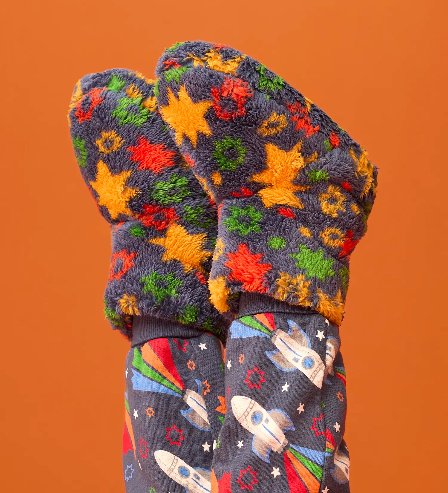 A pair of fuzzy knit mittens with a colorful star pattern in orange, yellow, green, and red on a dark gray background, shown on a child's hands paired with matching dark gray space-print leggings against a solid orange backdrop.