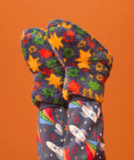 A pair of fuzzy knit mittens with a colorful star pattern in orange, yellow, green, and red on a dark gray background, shown on a child's hands paired with matching dark gray space-print leggings against a solid orange backdrop.