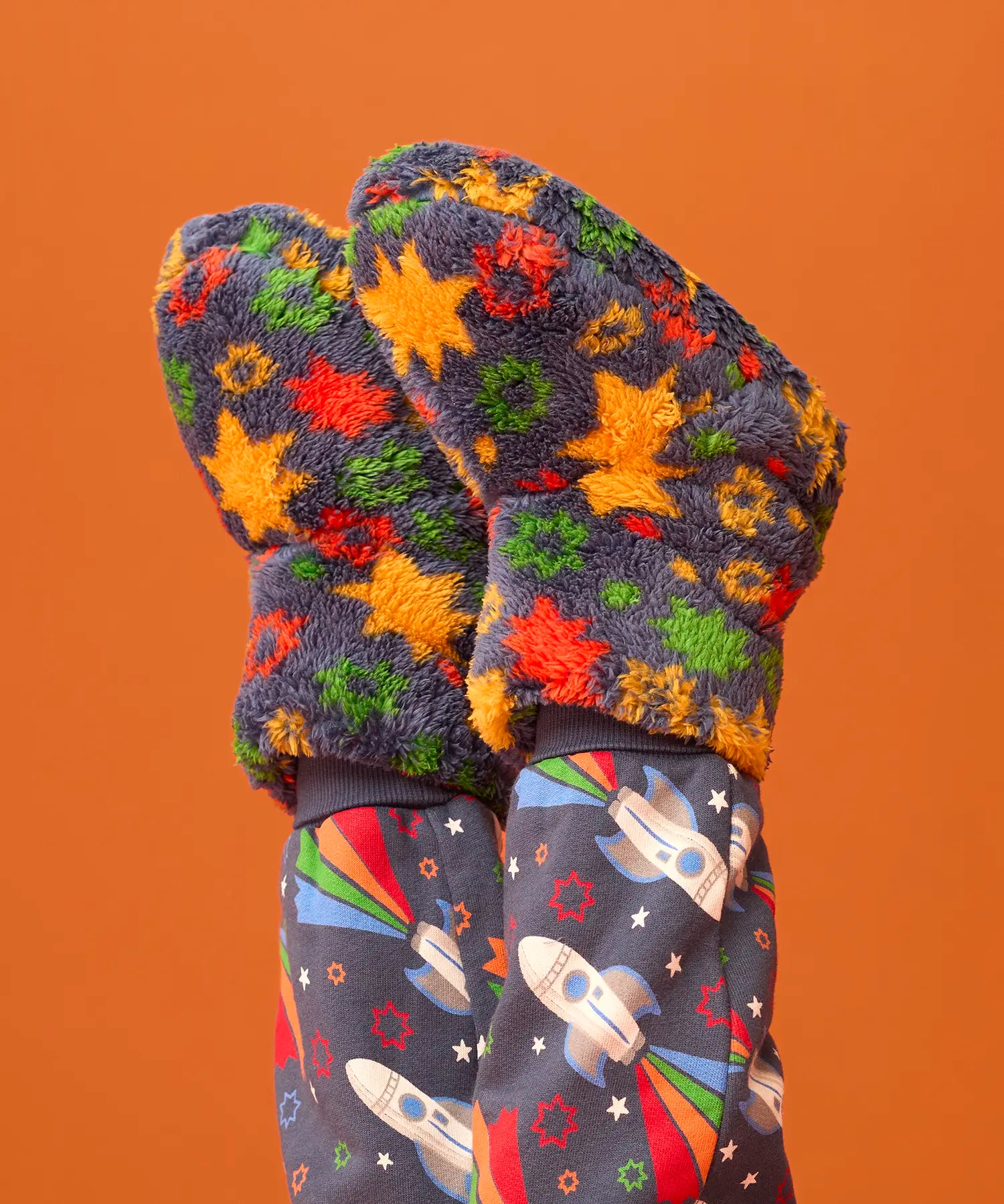 A pair of fuzzy knit mittens with a colorful star pattern in orange, yellow, green, and red on a dark gray background, shown on a child's hands paired with matching dark gray space-print leggings against a solid orange backdrop.