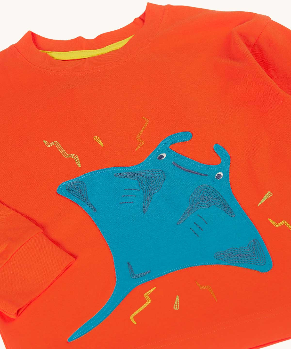 A close up of the front of the Frugi orange children's jumper with a large blue coloured ray applique on the front. 
