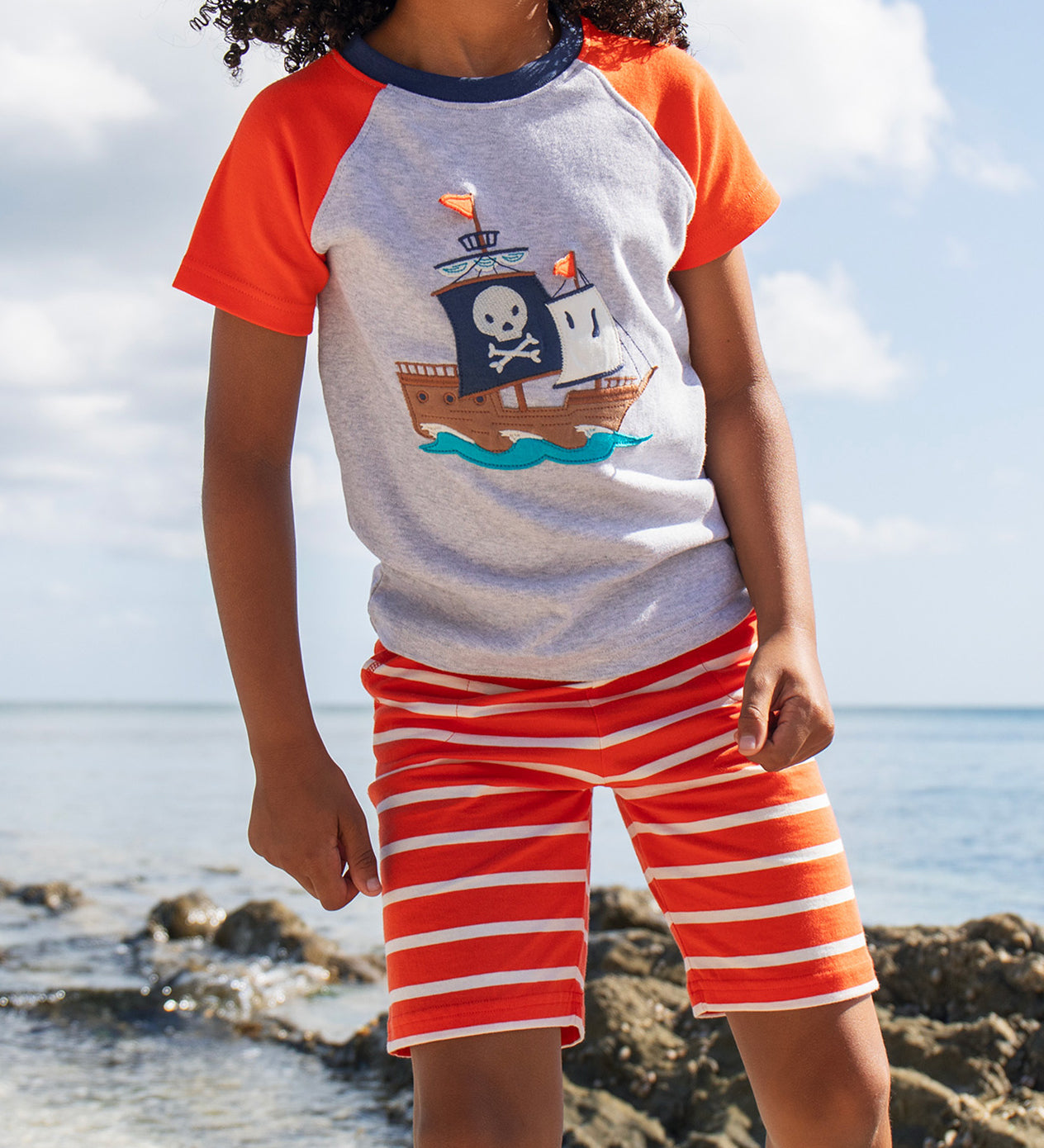 A young child wearing Frugi Stithian Shorts in Tiger Orange Breton, standing on a rock by the sea.