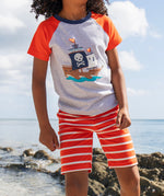 A young child wearing Frugi Stithian Shorts in Tiger Orange Breton, standing on a rock by the sea.