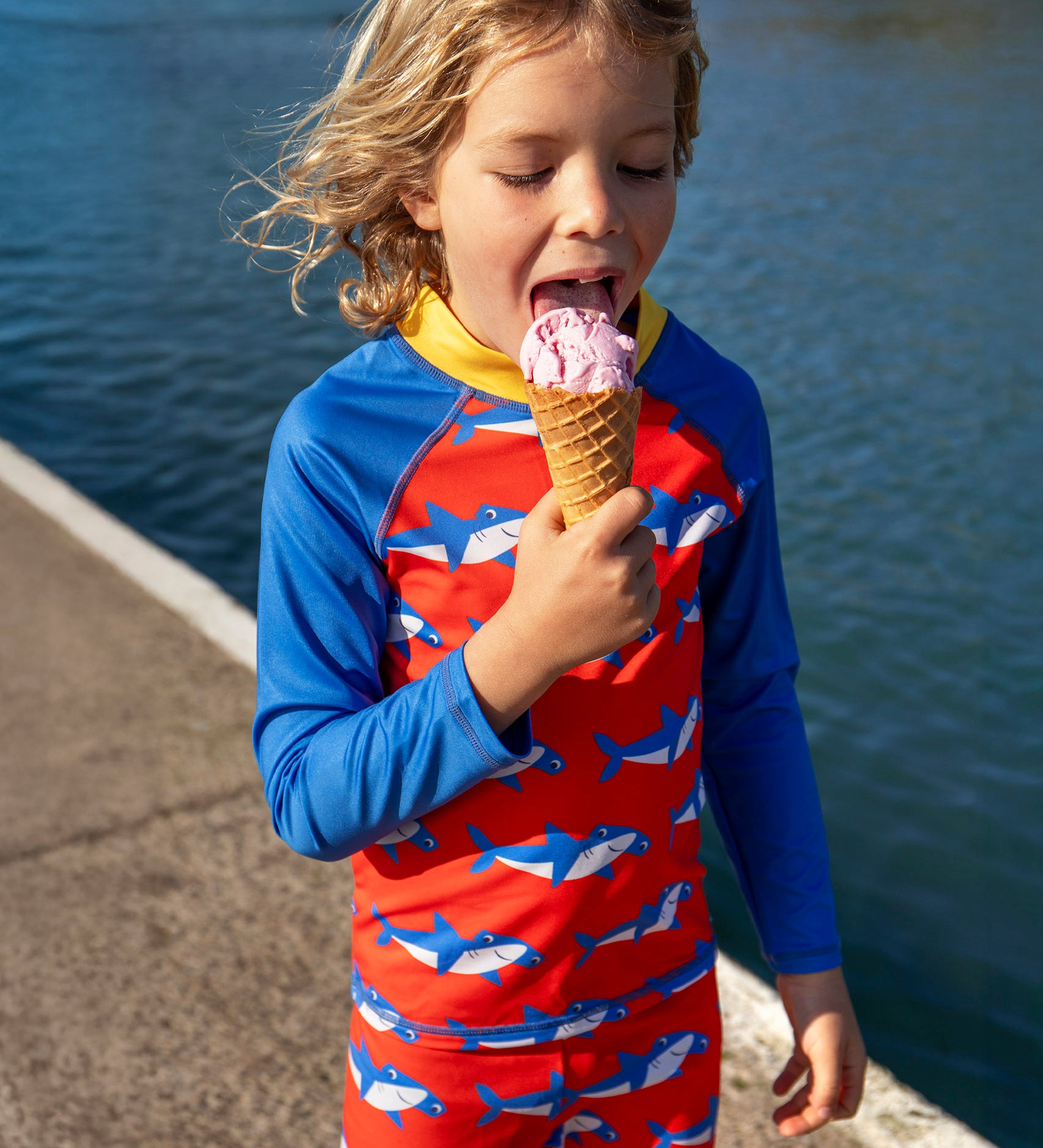Child wearing Frugi's happy sun safe swimwear set. Shows blue sleeves and red body with repeating hammerhead sharks designs