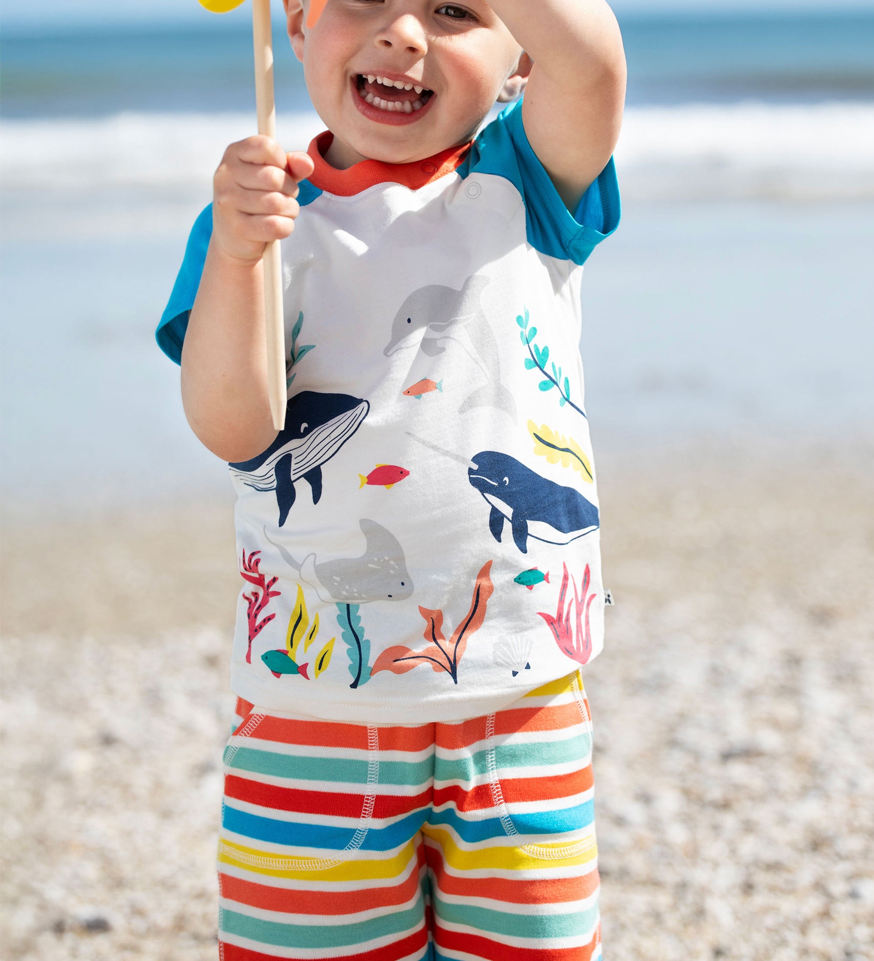 A smiling toddler wearing the Frugi Sunny Shorts in a Sea Rainbow Stripe print, standing on a pebbled beach holding a yellow pinwheel. 