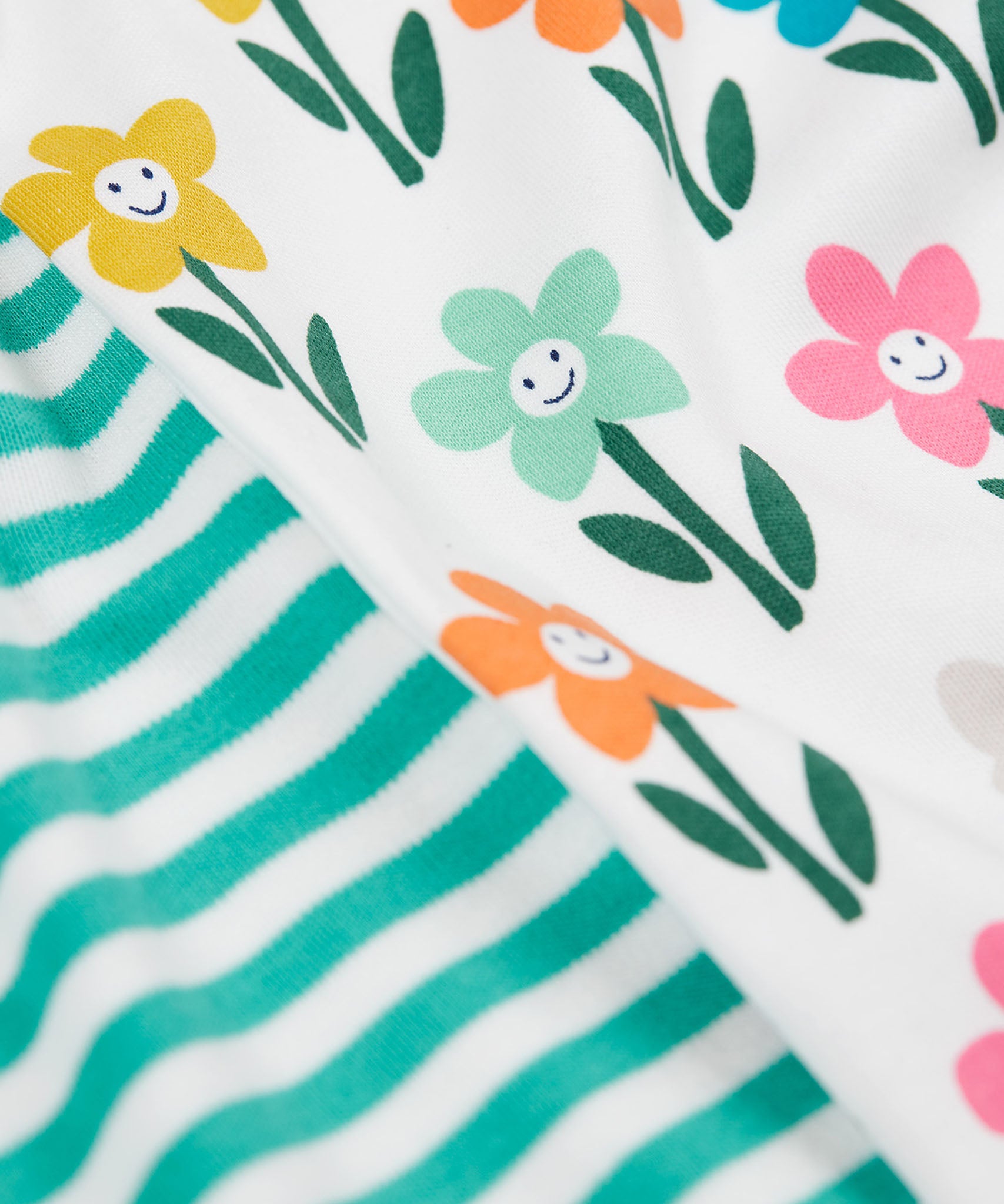 A super close up view of a set of two Frugi super special long sleeved organic cotton baby vests. One vest features the happy flowers print and the other has a turquoise and white striped design. 