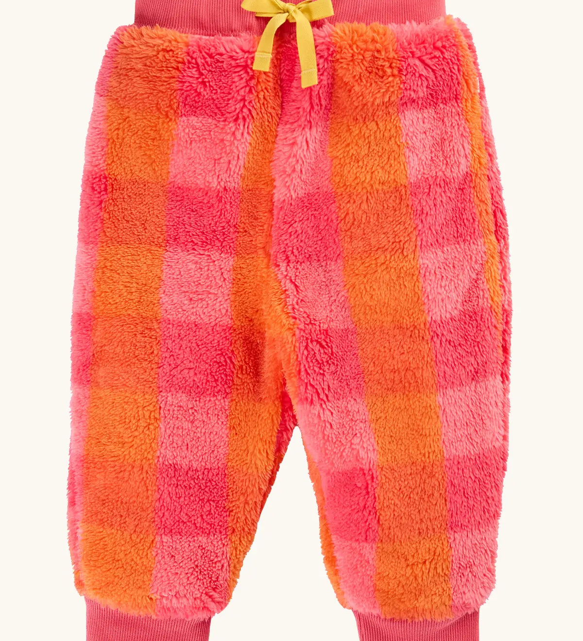A pair of frugi fuzzy, plush jogger-style pants in a pink and orange plaid pattern, featuring a yellow drawstring bow at the waist and ribbed cuffs at the ankles.