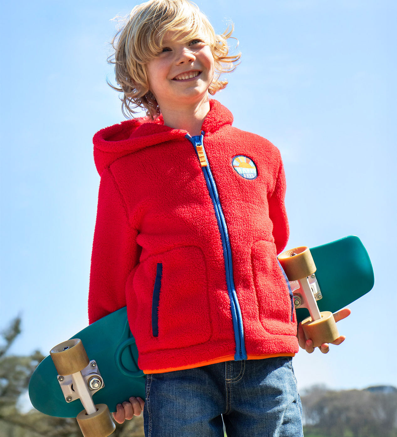 A boy with shoulder length blonde hair holding a blue skateboard behind his back wearing a Frugi red fleece hooded children's jacket. 