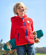 A boy with shoulder length blonde hair holding a blue skateboard behind his back wearing a Frugi red fleece hooded children's jacket. 