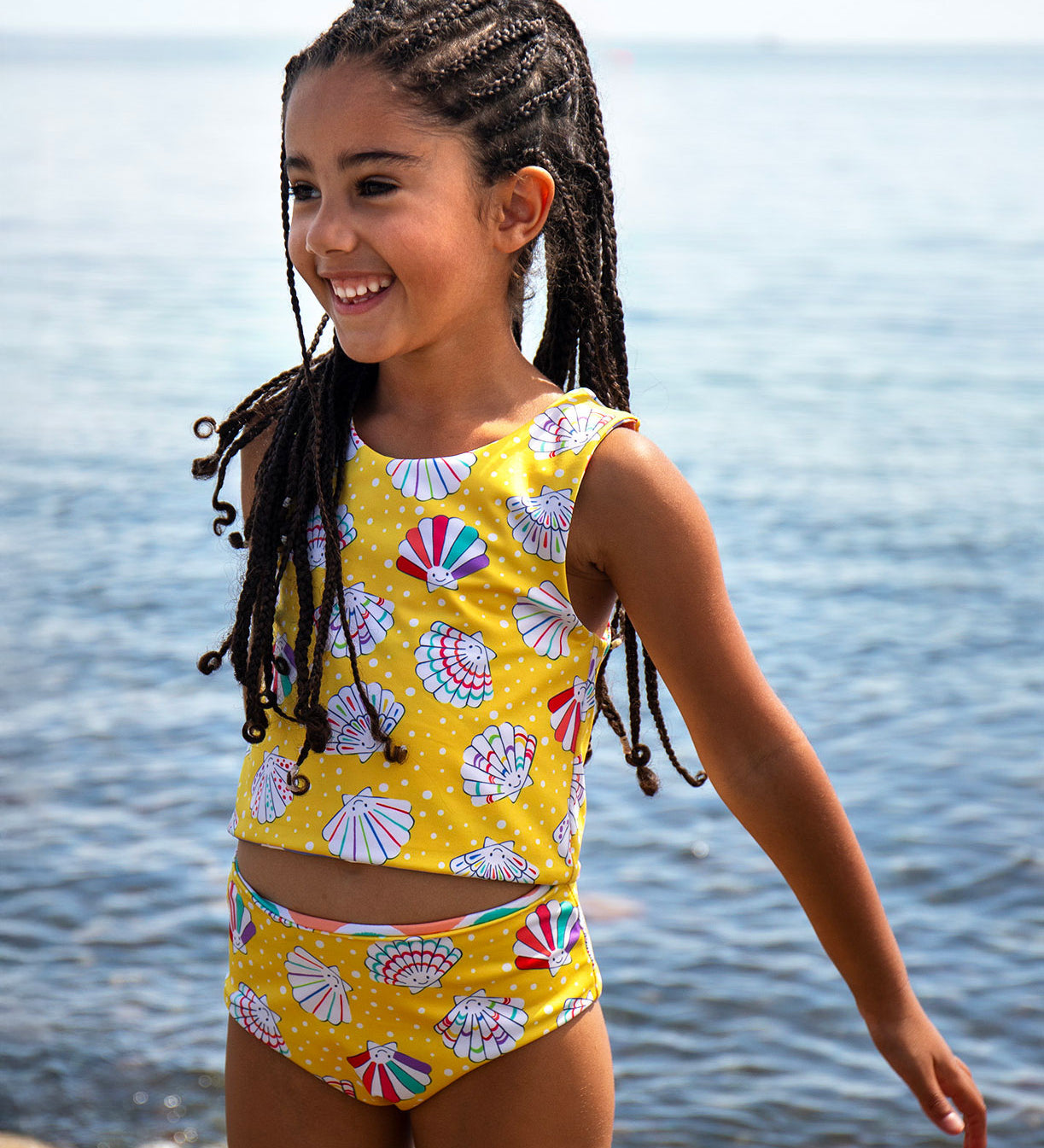 A girl with long dark braided hair standing in the sea wearing the Frugi reversible tankini with the yellow spotty side with shells all over showing. 