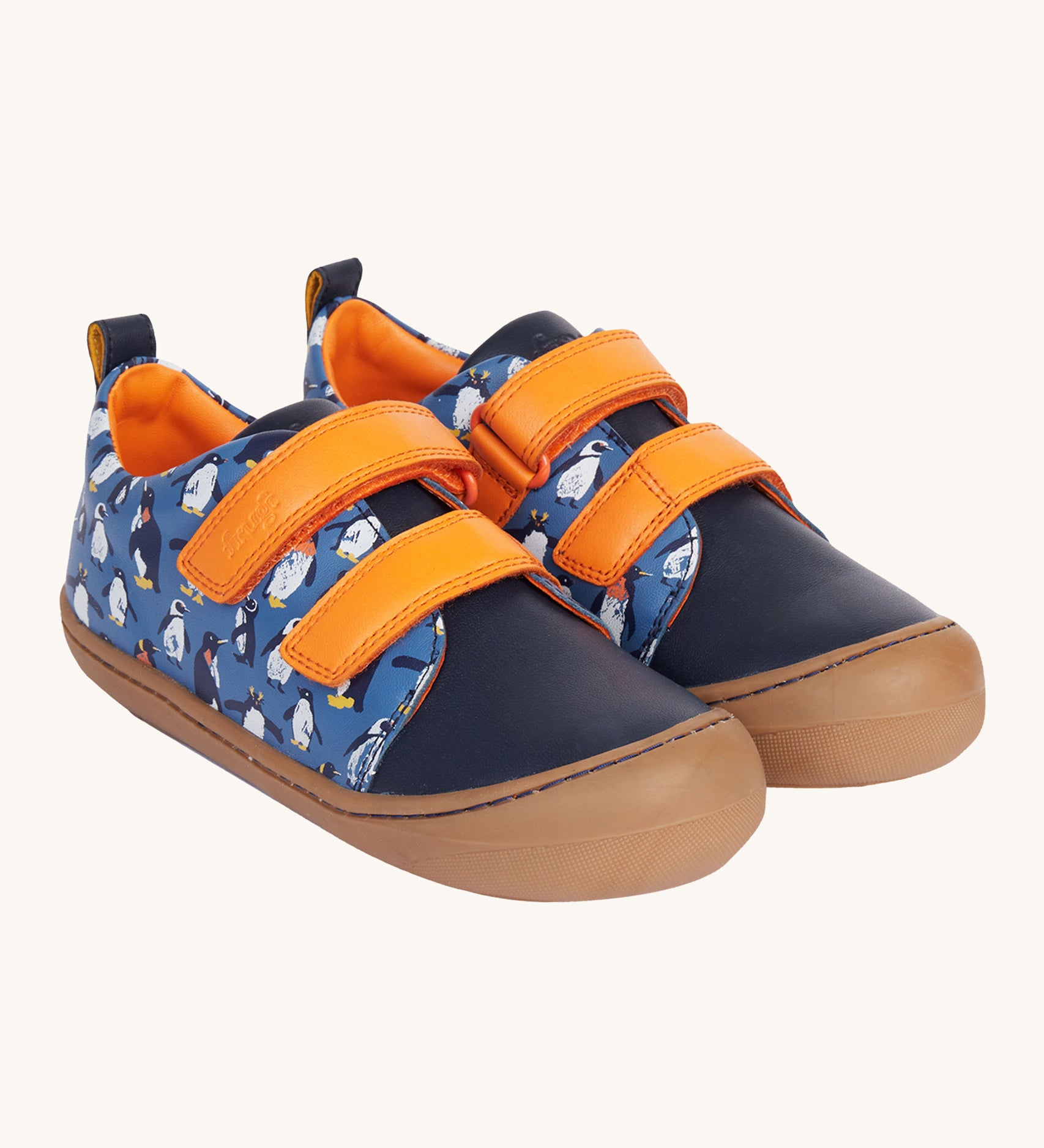 Frugi Penguin Print Tide Low Top velcro closure leather children's shoes. The side and back panels have a blue background with an all over penguin print, the front panel is a solid navy blue colour with contrasting orange coloured straps and lining. 