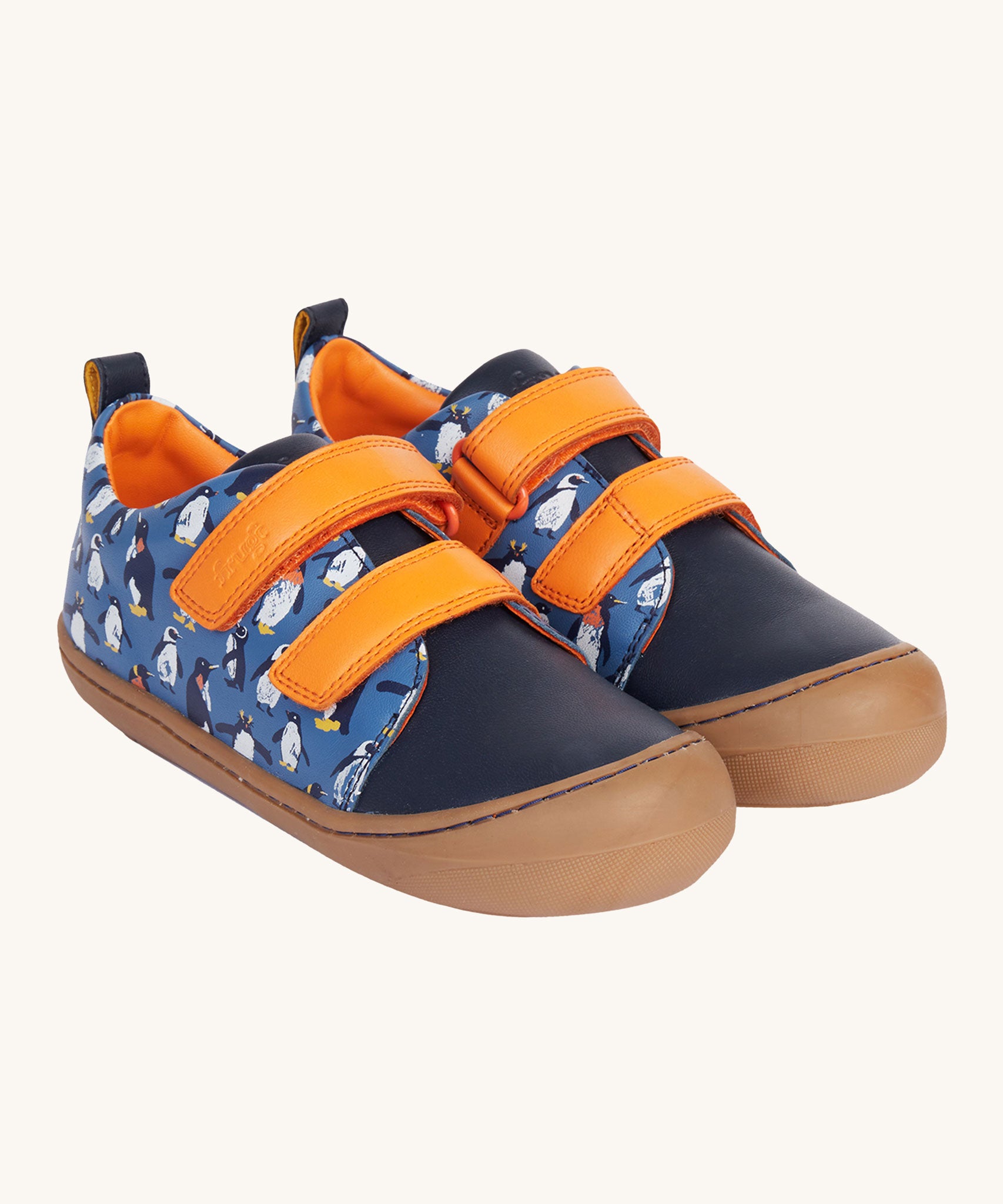 Frugi Penguin Print Tide Low Top velcro closure leather children's shoes. The side and back panels have a blue background with an all over penguin print, the front panel is a solid navy blue colour with contrasting orange coloured straps and lining. 