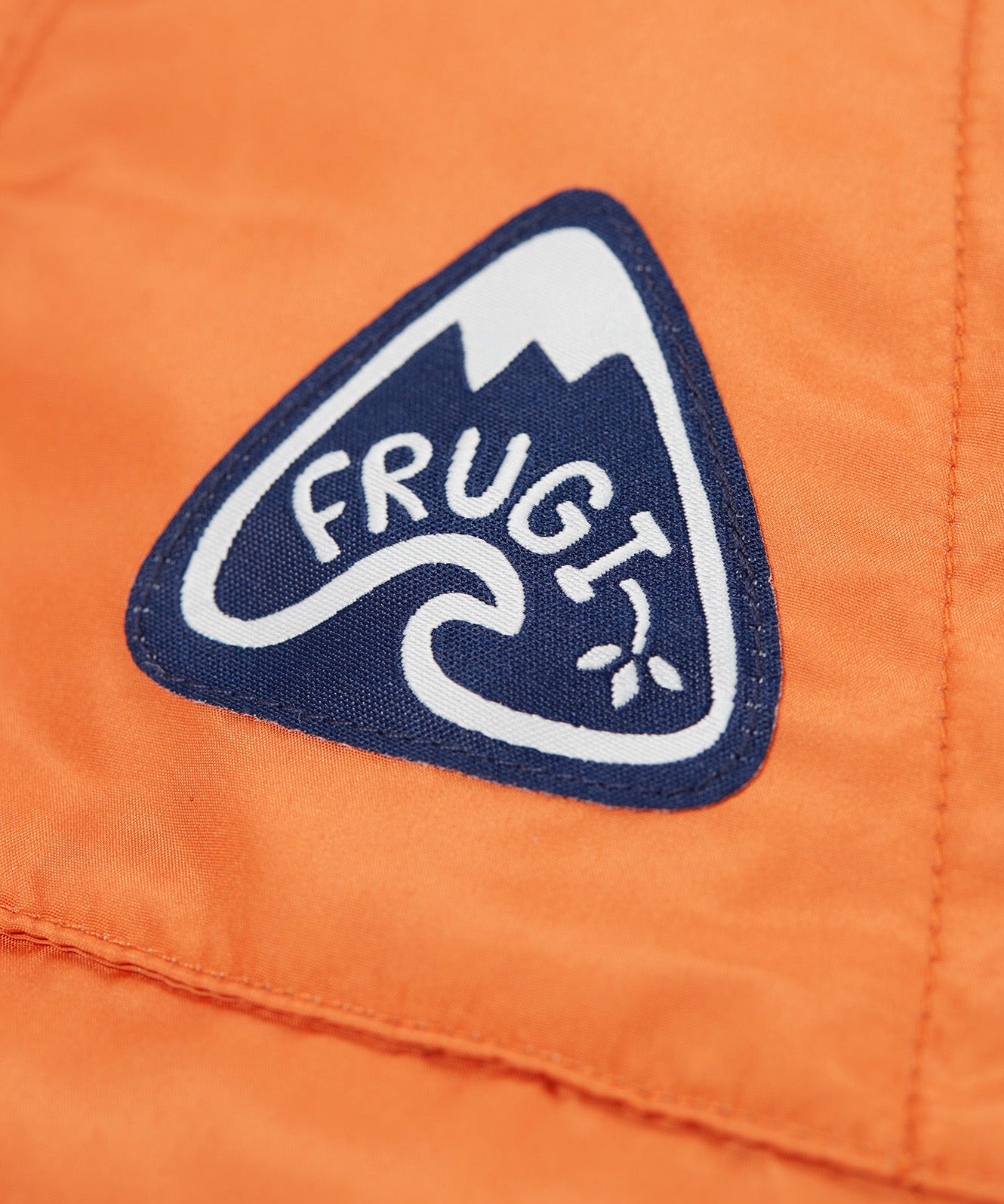 A close up of the Frugi toasty trail reversible jacket showing the brand patch stitched on the side pocket on orange post consumer recycled polyester fabric