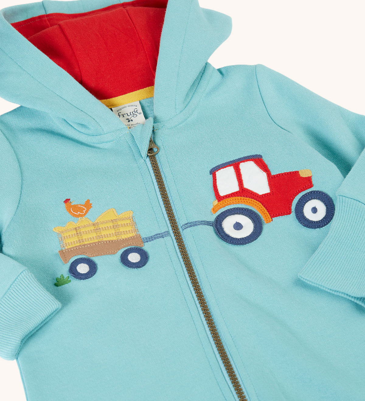 A close up of the front of the Frugi soft green all in one organic cotton zip suit showing the tractor and trailer applique and red lined hood.