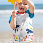 A smiling toddler wearing the Frugi Reid Raglan T-Shirt in Soft White, featuring a Sea Life Whale print, standing on a pebbled beach and holding a yellow pinwheel.
