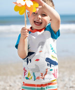 A smiling toddler wearing the Frugi Reid Raglan T-Shirt in Soft White, featuring a Sea Life Whale print, standing on a pebbled beach and holding a yellow pinwheel.