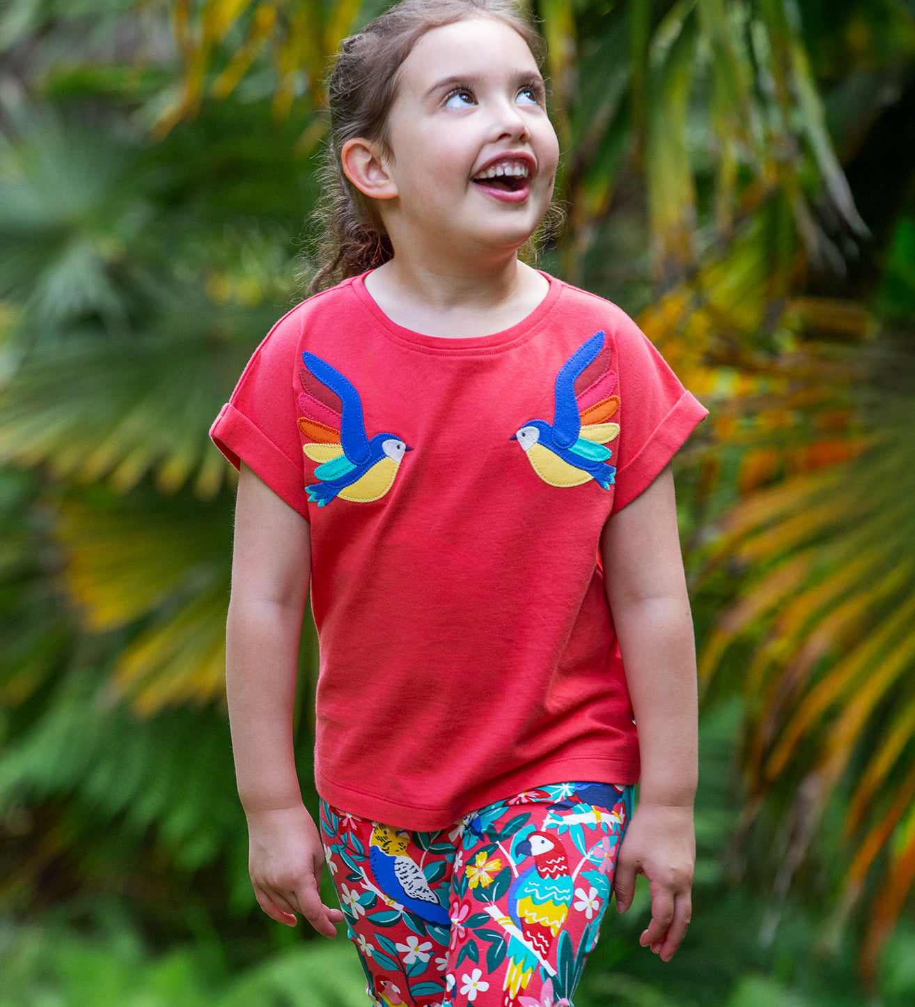 A smiling toddler wearing the Frugi Daisy Applique T-Shirt in Watermelon, featuring two embroidered parrots on either side, walking amongst exotic plants.
