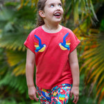 A smiling toddler wearing the Frugi Daisy Applique T-Shirt in Watermelon, featuring two embroidered parrots on either side, walking amongst exotic plants.