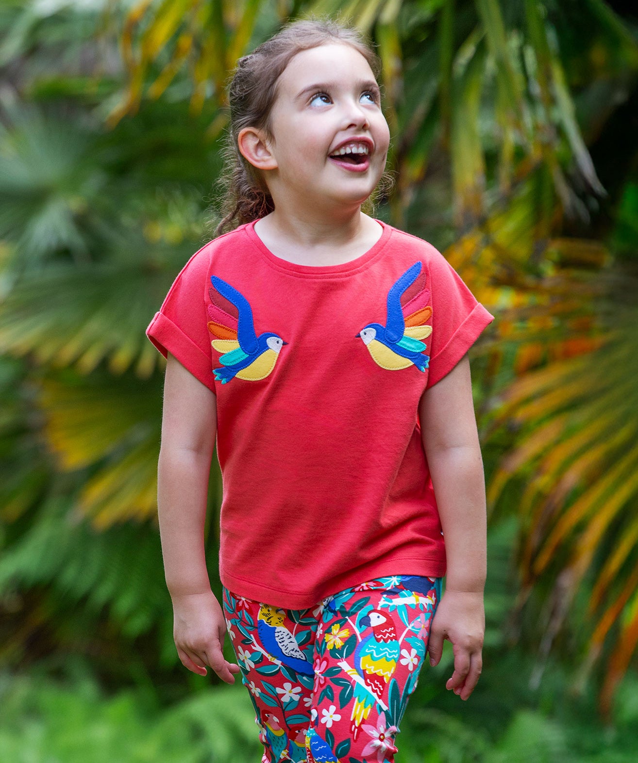 A smiling toddler wearing the Frugi Daisy Applique T-Shirt in Watermelon, featuring two embroidered parrots on either side, walking amongst exotic plants.