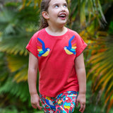 A smiling toddler wearing the Frugi Daisy Applique T-Shirt in Watermelon, featuring two embroidered parrots on either side, walking amongst exotic plants.