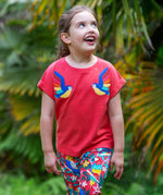 A smiling toddler wearing the Frugi Daisy Applique T-Shirt in Watermelon, featuring two embroidered parrots on either side, walking amongst exotic plants.