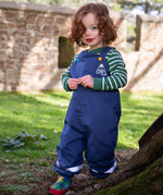 A toddler standing outside wearing the Frugi navy waterproof dungarees with reflective straps on the legs. Features a blue and yellow button fastener for the shoulder strap for a secure fit