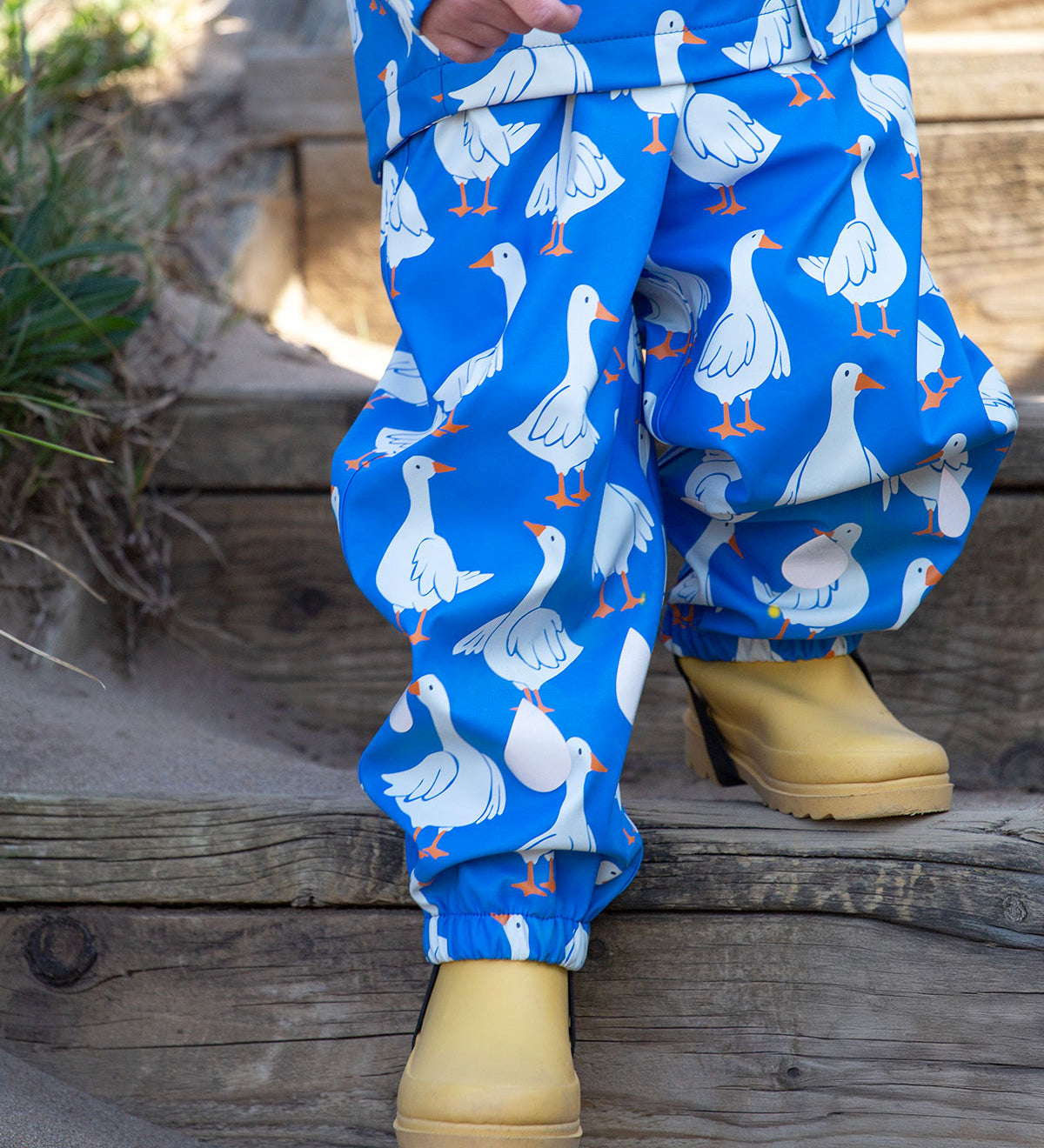 A close up of a toddler's legs walking down sandy steps on a beach wearing the Frugi blue goose print puddle buster trousers with yellow welly boots. 