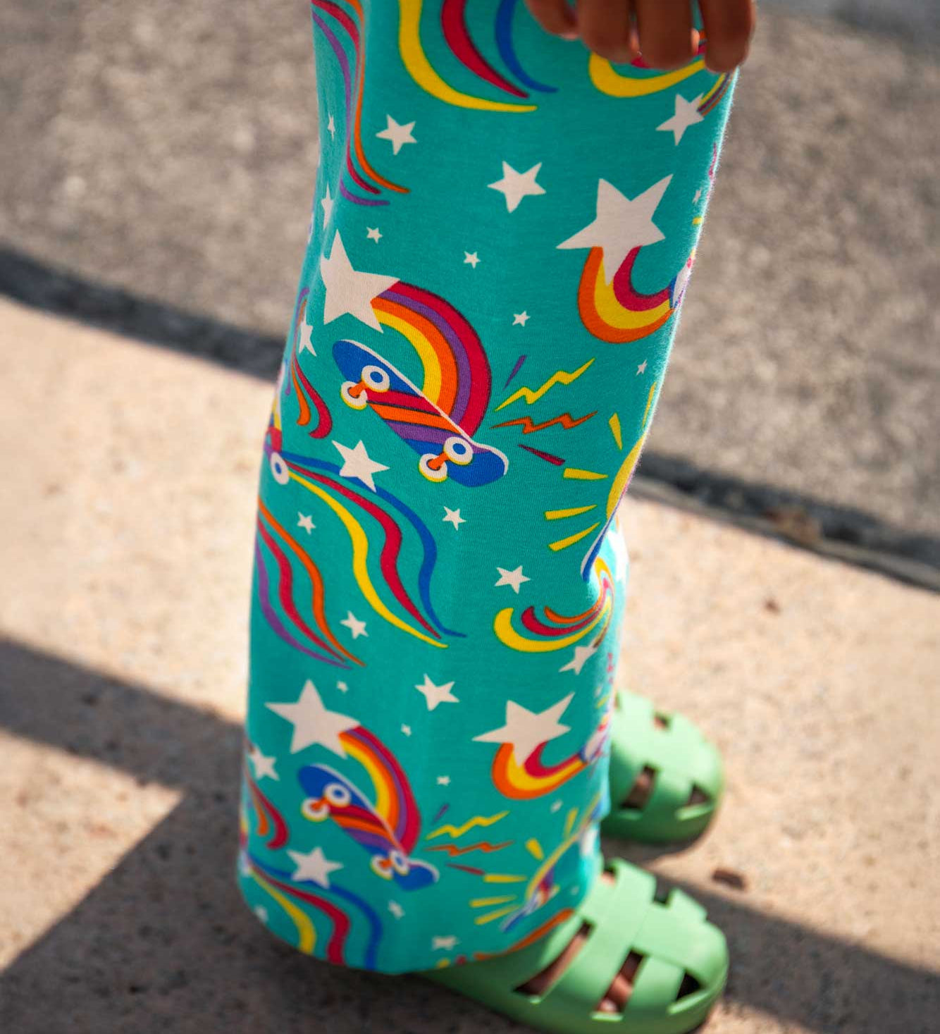 A close up of a child's legs wearing the Frugi wide leg trousers with an all over print on a teal background featuring colourful skateboards and roller blades with green colured jelly shoes. 