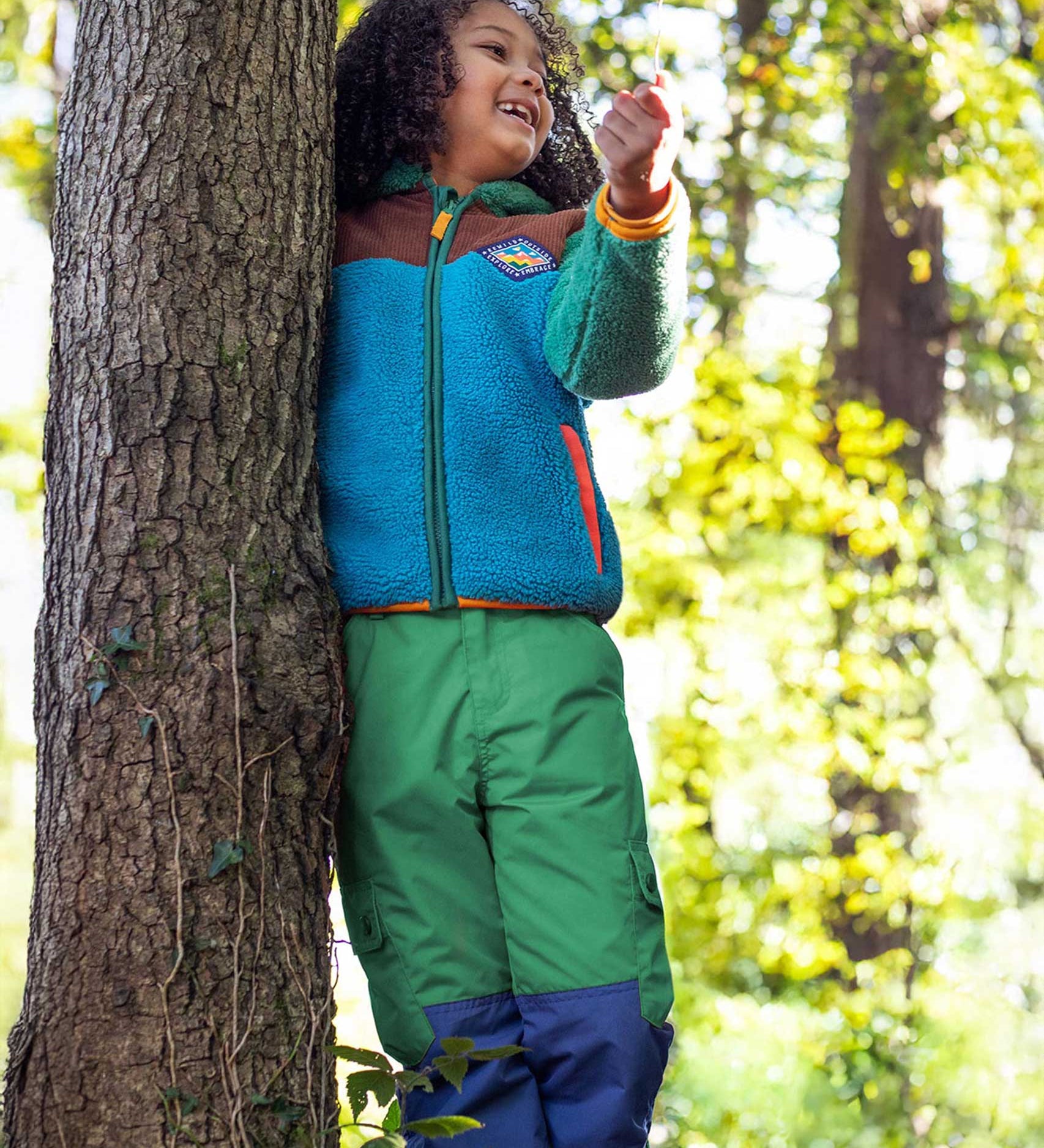 A child wearing the Frugi Wilderness green trousers with navy blue coloured knee patches with a Frugi colour block fleece. 