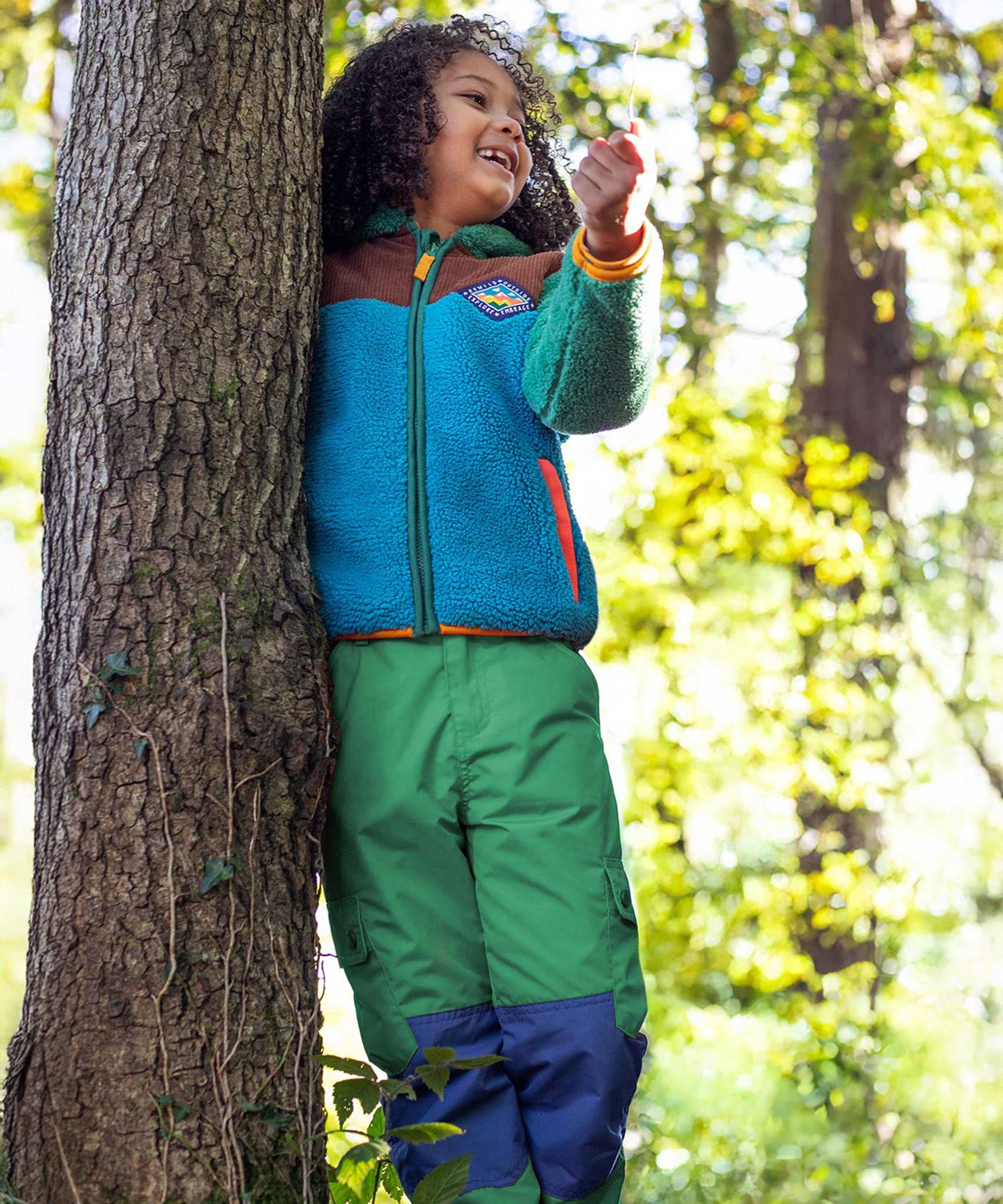 A child wearing the Frugi Wilderness green trousers with navy blue coloured knee patches with a Frugi colour block fleece. 