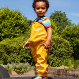 Child wearing the Frugi children's waterproof Puddle buster trousers with elasticated arm straps, in bright Bumble Bee Yellow with a rainbow striped t-shirt underneath.