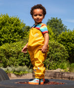 Child wearing the Frugi children's waterproof Puddle buster trousers with elasticated arm straps, in bright Bumble Bee Yellow with a rainbow striped t-shirt underneath.