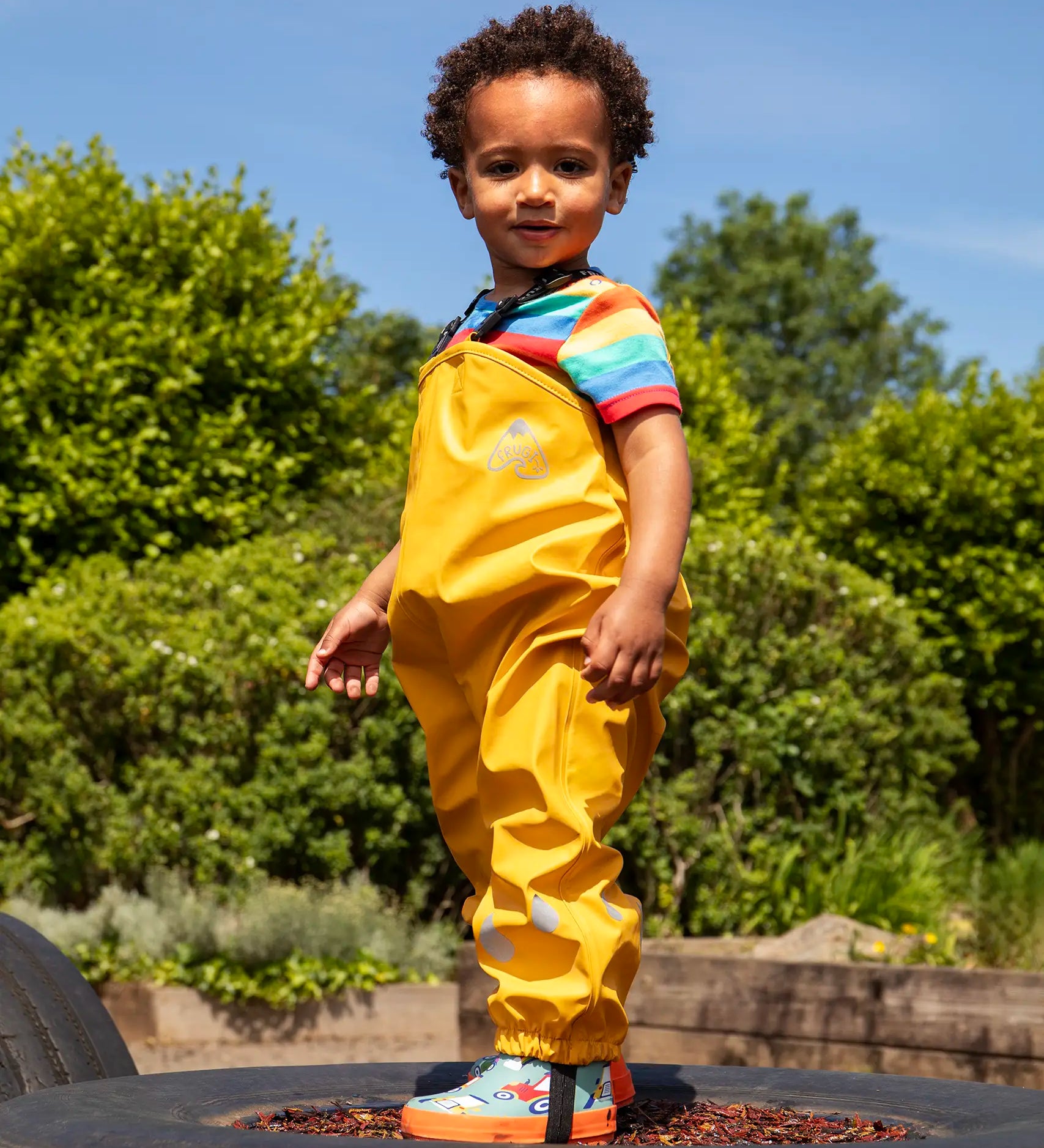 Child wearing the Frugi children's waterproof Puddle buster trousers with elasticated arm straps, in bright Bumble Bee Yellow with a rainbow striped t-shirt underneath.