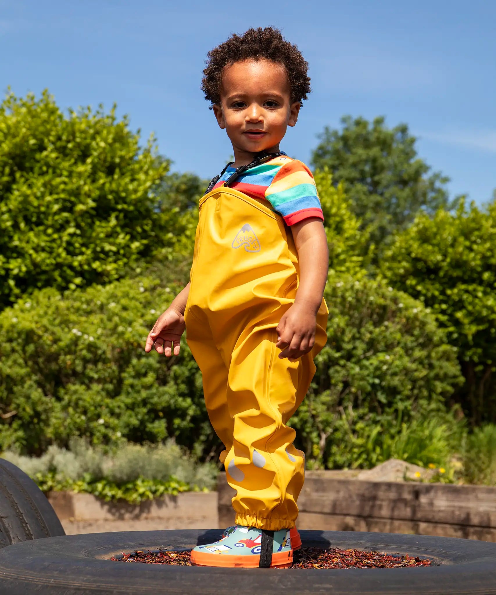 Child wearing the Frugi children's waterproof Puddle buster trousers with elasticated arm straps, in bright Bumble Bee Yellow with a rainbow striped t-shirt underneath.